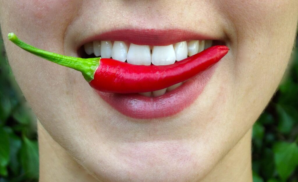 Eine lächelnde Person mit weißen Zähnen hält eine rote Chilischote zwischen den Lippen – trotz der Gefahr von Entzündungen im Mund. Der Hintergrund ist verschwommenes Grün.